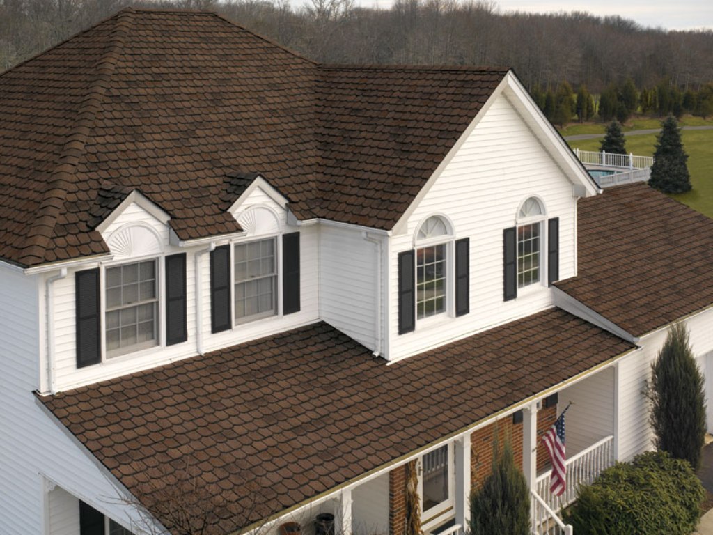 A two-story house with a white exterior and dark brown architectural shingles roof. The house features multiple dormer windows with black shutters, a front porch with white columns, and an American flag. Trees and a lawn surround the property.