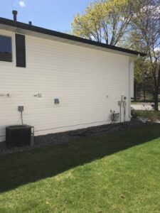 White exterior wall of a house with several utility meters and an air conditioning unit on a gravel bed. Lush green grass in the foreground and a clear blue sky with trees in the background.