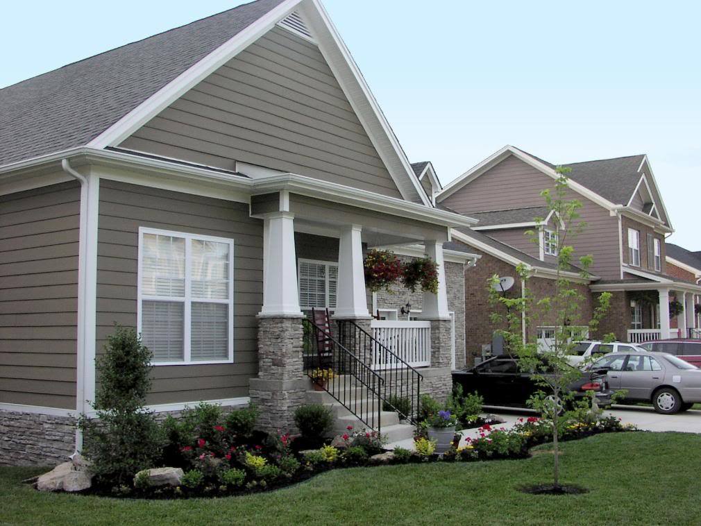 A small, charming suburban house with beige siding and a white trim. It has a covered front porch with columns, a manicured lawn, colorful flower beds, and a young tree in front. Two neighboring houses and cars parked in the driveway are visible.