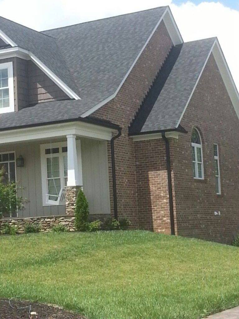 A two-story brick house with white trim and dark shingles sits on a well-maintained lawn with a small tree and shrubbery. The house has a gabled roof, a covered front porch supported by white columns, and several windows including an arched one on the right side.