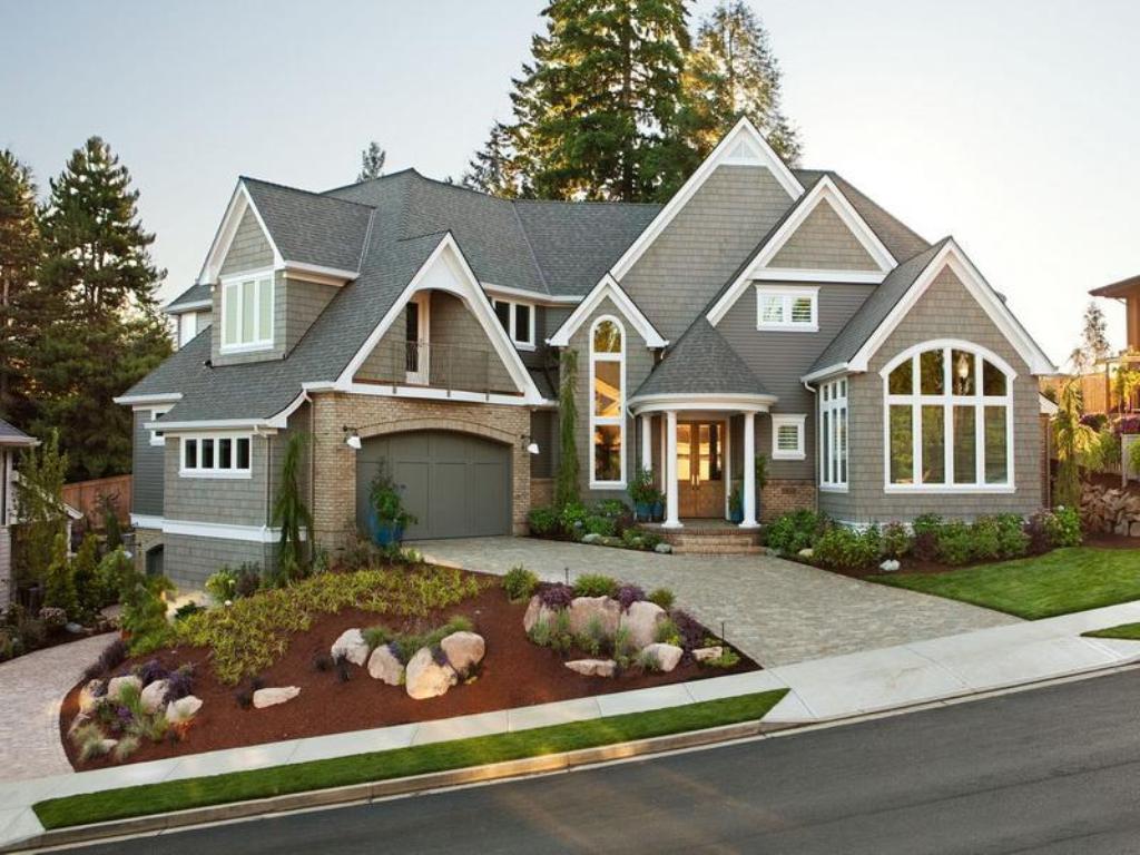 A large, modern suburban house with a gabled roof, a three-car garage, and a neatly manicured lawn. The exterior features gray shingles, large windows, and a mix of stone and wooden elements. Trees and shrubs are arranged in the front yard, with a paved driveway and walkway.