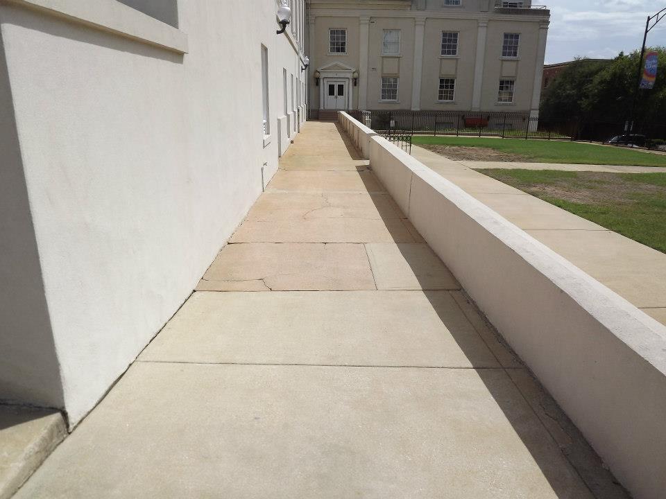 A concrete ramp beside a building leads to an access door, perfect for easy patio cleaning services access. The ramp has a handrail on one side and a retaining wall on the other, with a grassy area visible in the background. The sky is clear, casting shadows along the path.