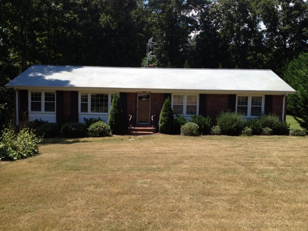 A single-story brick house with large white-framed windows, a central wooden door, and a light-colored roof. The front yard has well-maintained bushes and a large grassy lawn. Surrounded by trees, this home shines after professional Soft Wash services that bring out its true charm.