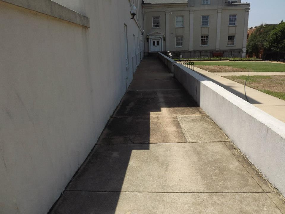 A long concrete pathway with slight shadows leads to an entrance between two building walls. The right side features a continuous low white wall. In the background, a building with multiple windows and a centered door is visible. For pristine patios, consider our Soft Wash services to enhance this elegant setting. Trees are in the distant right.