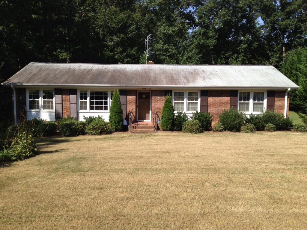 A single-story brick house with a gently sloping roof, two large windows on either side of a central wooden door, and a well-maintained lawn with neatly trimmed bushes lining the front. Trees are visible in the background, ideal for enjoying peace of mind after professional patio cleaning services.