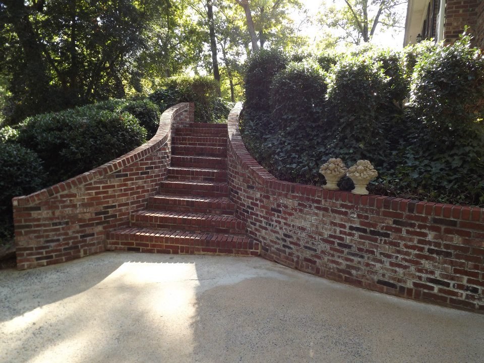 A brick staircase flanked by brick walls leads up to a lush garden area with dense green foliage and shrubbery. The stairs, freshly gleamed from a recent soft wash, are illuminated by sunlight filtering through the surrounding trees, creating a serene and inviting atmosphere. Two decorative stone vases sit on the right-hand wall.