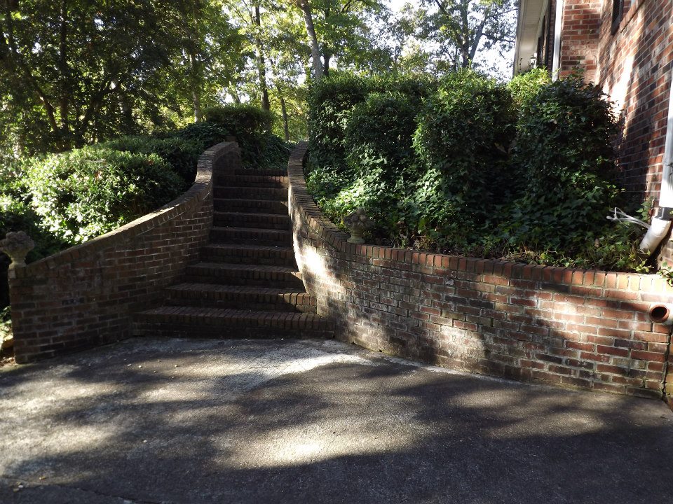 A flight of brick steps leads up between two curved brick walls, surrounded by greenery and bushes, to a higher garden level next to a brick house. The area, maintained with our patio cleaning services and shaded by tall trees, creates a serene and inviting outdoor atmosphere.