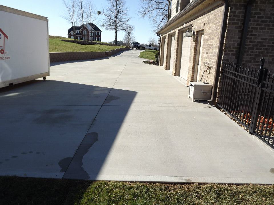 A long, newly paved concrete driveway leading to a house with a three-car garage. A white moving container is parked on the left side. The driveway, perfect for showcasing our soft wash patio cleaning services, is bordered by a green lawn and a wrought-iron fence on the right. A large house is visible in the background.