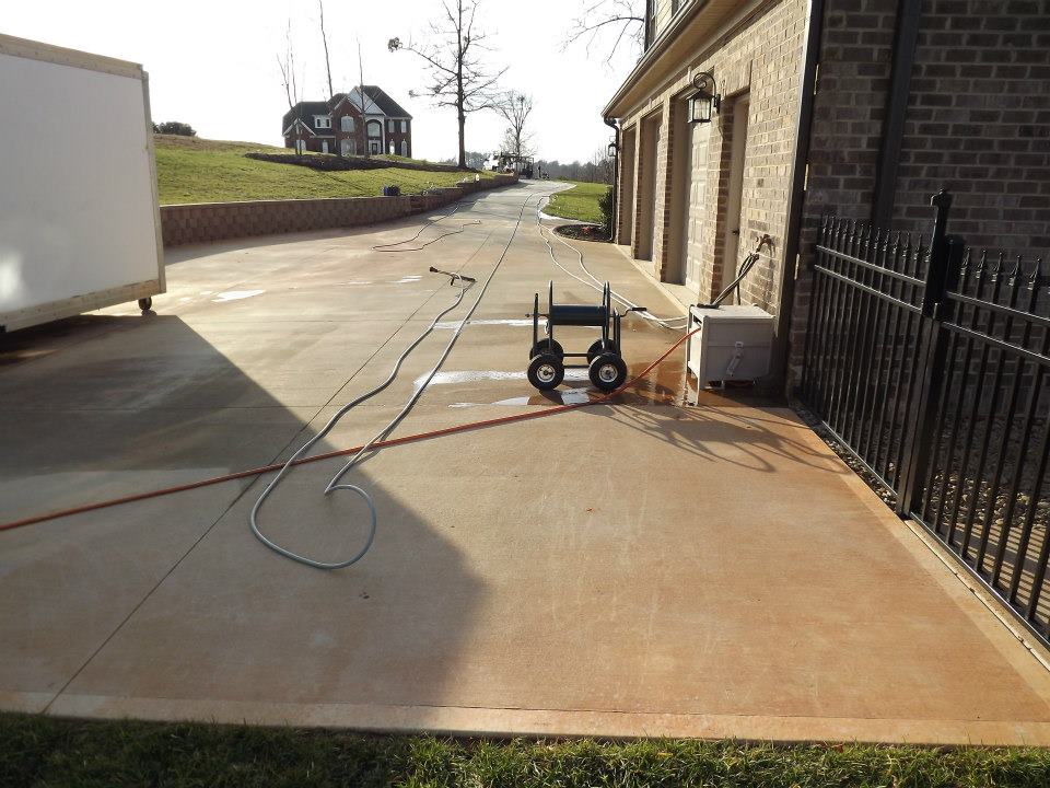 A clean, wide driveway leads to a brick house. Near the garage, equipment like hoses and a pressure washer are set up, possibly for patio cleaning or soft wash services. A large white container is on the left, and a black metal fence is on the right.