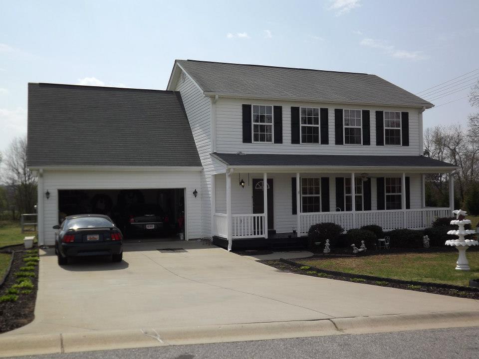 A two-story white house with black shutters and a wraparound porch stands elegantly. The attached garage on the left side houses a sleek black car. The smooth driveway extends to the street, bordered by a manicured lawn with small bushes and a decorative fountain. Expert soft wash services keep it pristine.