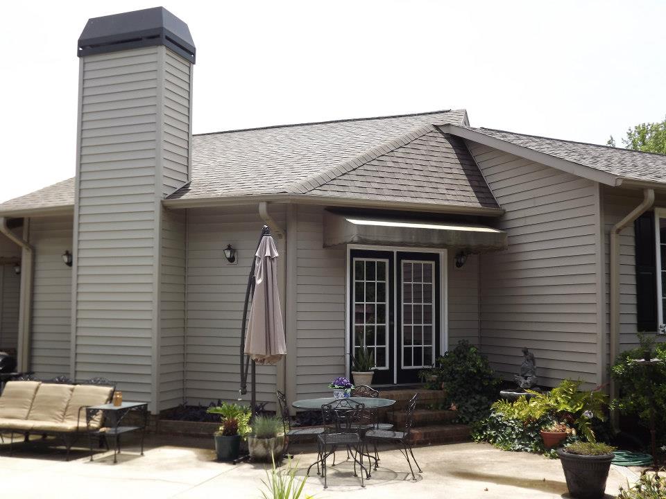 A backyard patio features a beige house with gray trim, double French doors, and a porch light. Outdoor furnishings include a metal table with chairs, a bench with cushions, an umbrella, and various potted plants. The house has a steeply pitched roof and chimney; perfect for Soft Wash patio cleaning services.