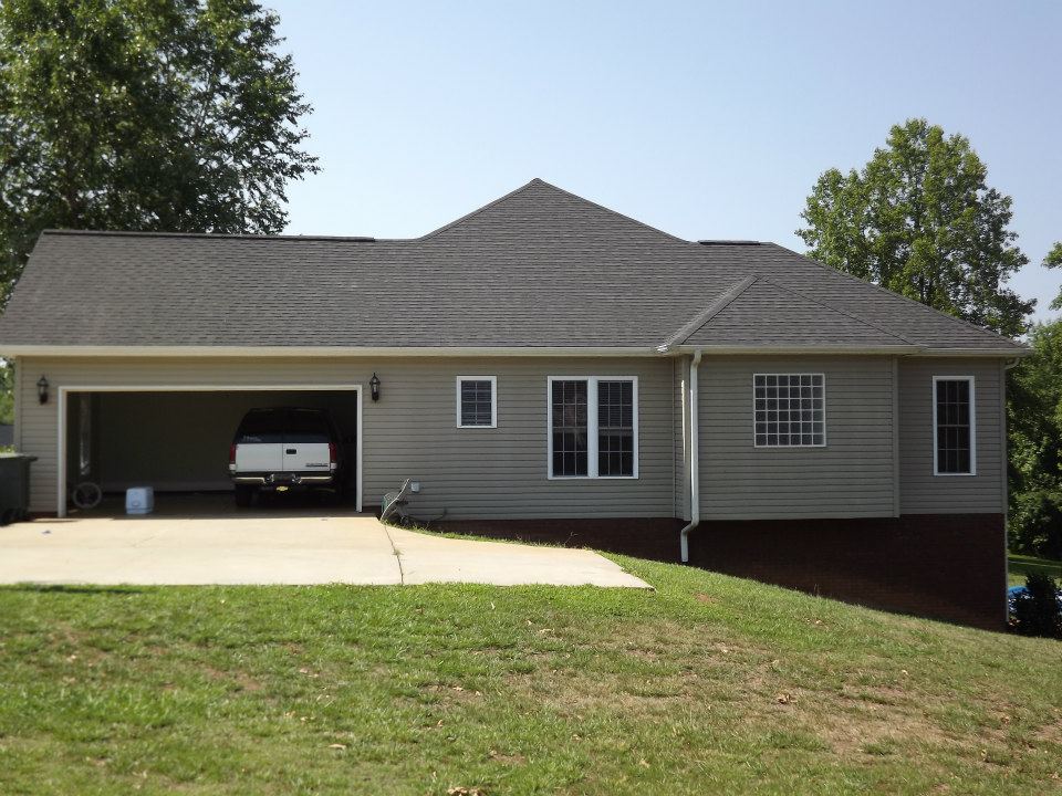 A single-story house with a gray exterior and a dark gray roof is shown. The garage, partially open, contains a vehicle. The driveway leads up to the garage, and the front yard is grassy with a slight slope. The property benefits from expert patio cleaning services, enhancing its pristine look. Trees are visible in the background.