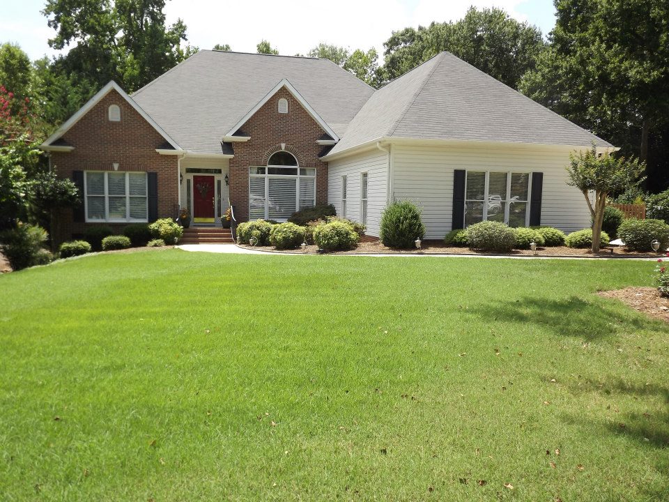 A single-story house with a brick façade and white siding, featuring a gabled roof and a well-manicured front lawn. The entryway is flanked by neatly trimmed bushes, and a large window above the entrance adds to the home's charm. Trees surround the property, offering privacy perfect for soft wash services.