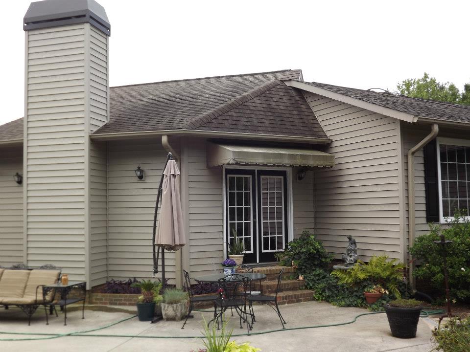 A cozy backyard scene featuring the exterior of a house with beige siding and a dark shingled roof. There's a chimney on the left, French doors with an awning in the center, and various potted plants and patio furniture, including chairs and a table—perfect for relaxing after a professional patio cleaning.