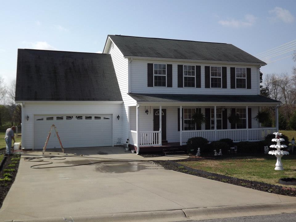 A two-story white house with black shutters and a green roof boasts a spacious attached garage, a large driveway, and a well-kept lawn. The covered front porch features white railings. A person is visible providing soft wash services near a ladder in the driveway. A decorative white bird bath sits on the lawn.