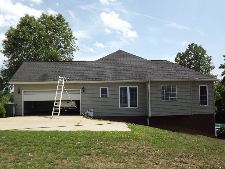 A single-story house with beige siding and a dark roof is shown from the front. A double garage door is open, and a white extension ladder leans against the roof. A garden hose, perfect for patio cleaning services or a soft wash, is coiled nearby on the concrete driveway. The background features green trees and a blue sky.