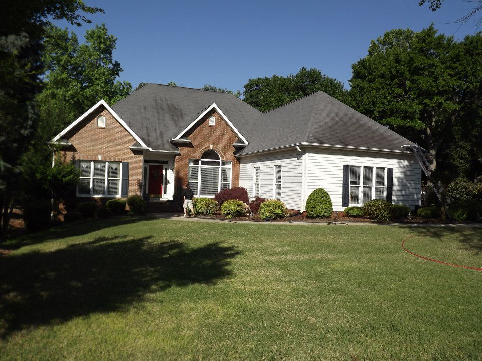 A single-story house with brick and white siding, featuring a steep gable roof and multiple windows, is situated in a well-manicured front yard. A person stands near the entrance on a sunny day, enjoying the freshly cleaned patio, thanks to expert soft wash services, with lush green trees in the background.