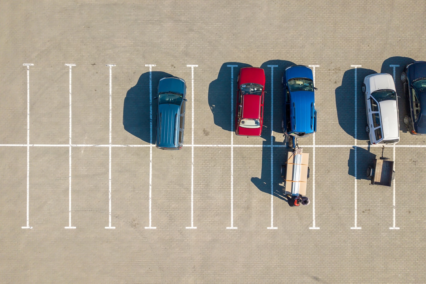 An aerial view of a nearly empty parking lot with seven parking spaces. Four vehicles are parked in the first four spaces from the left - a blue car, a red car, a blue SUV, and a silver car. Two people are loading a cart with wooden planks next to the blue SUV while another cart advertises pressure washing services.