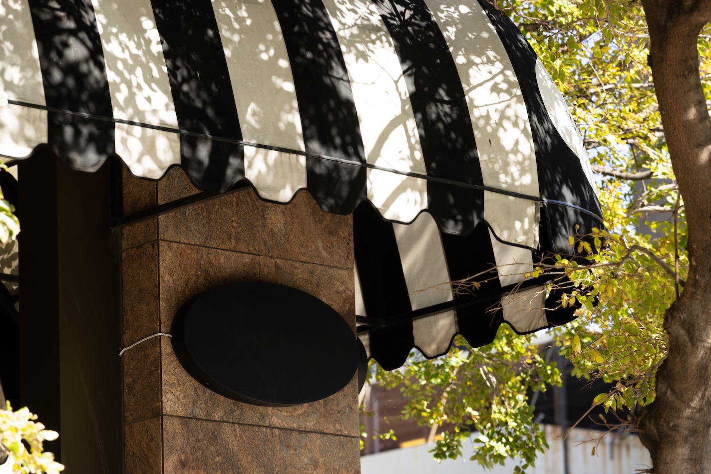 A black oval sign hanging on the brown stone exterior of a building with a black and white striped awning looks pristine, as if recently pressure washed. Sunlight filters through the leaves of nearby trees, casting shadows on the awning and sign.