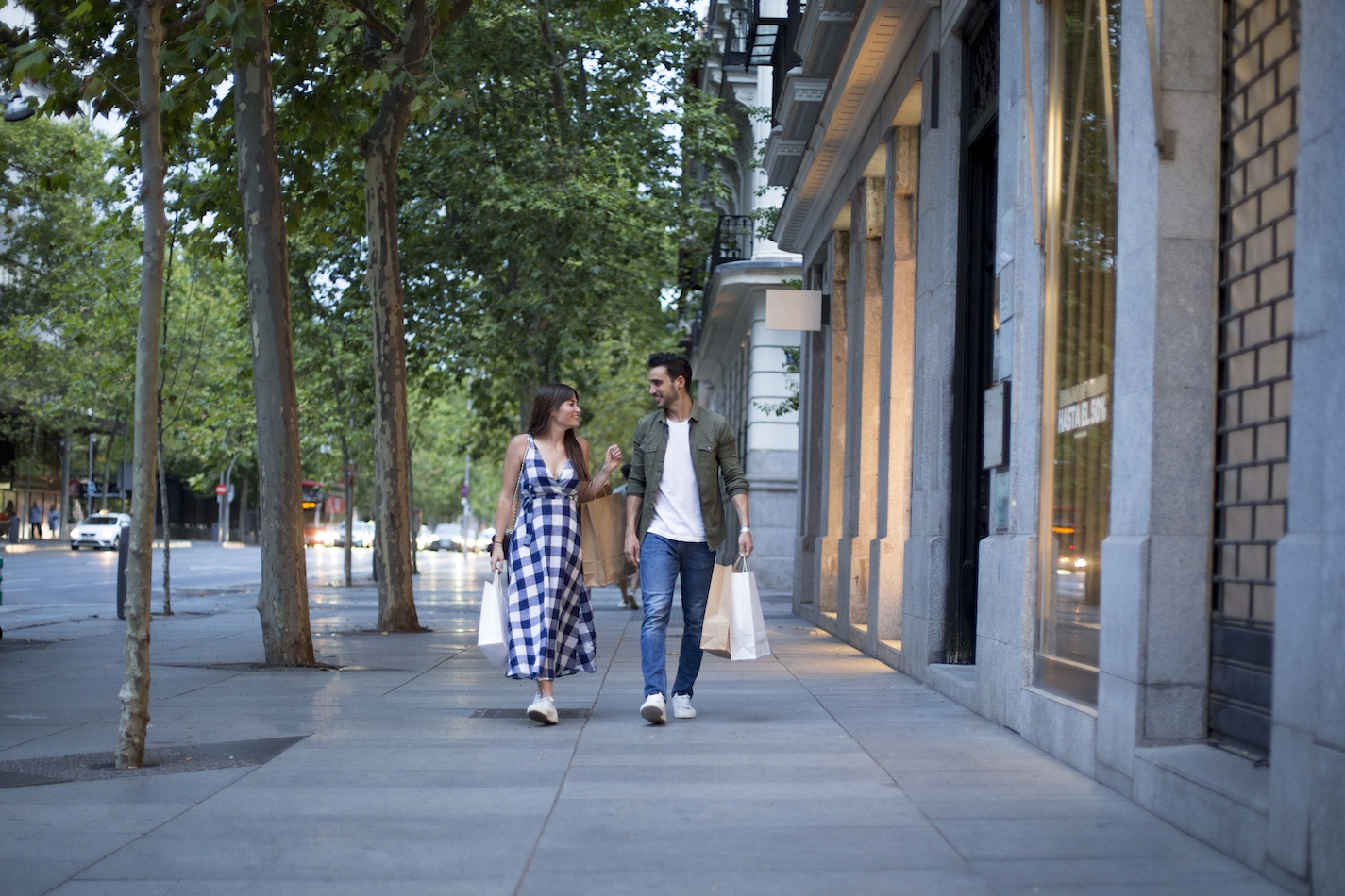 A woman in a blue and white checkered dress and a man in casual clothes walk along an urban street lined with trees and shops. Smiling at each other, they both hold shopping bags. In the background, a worker is pressure washing the sidewalk on this sunny day with light filtering through the trees.