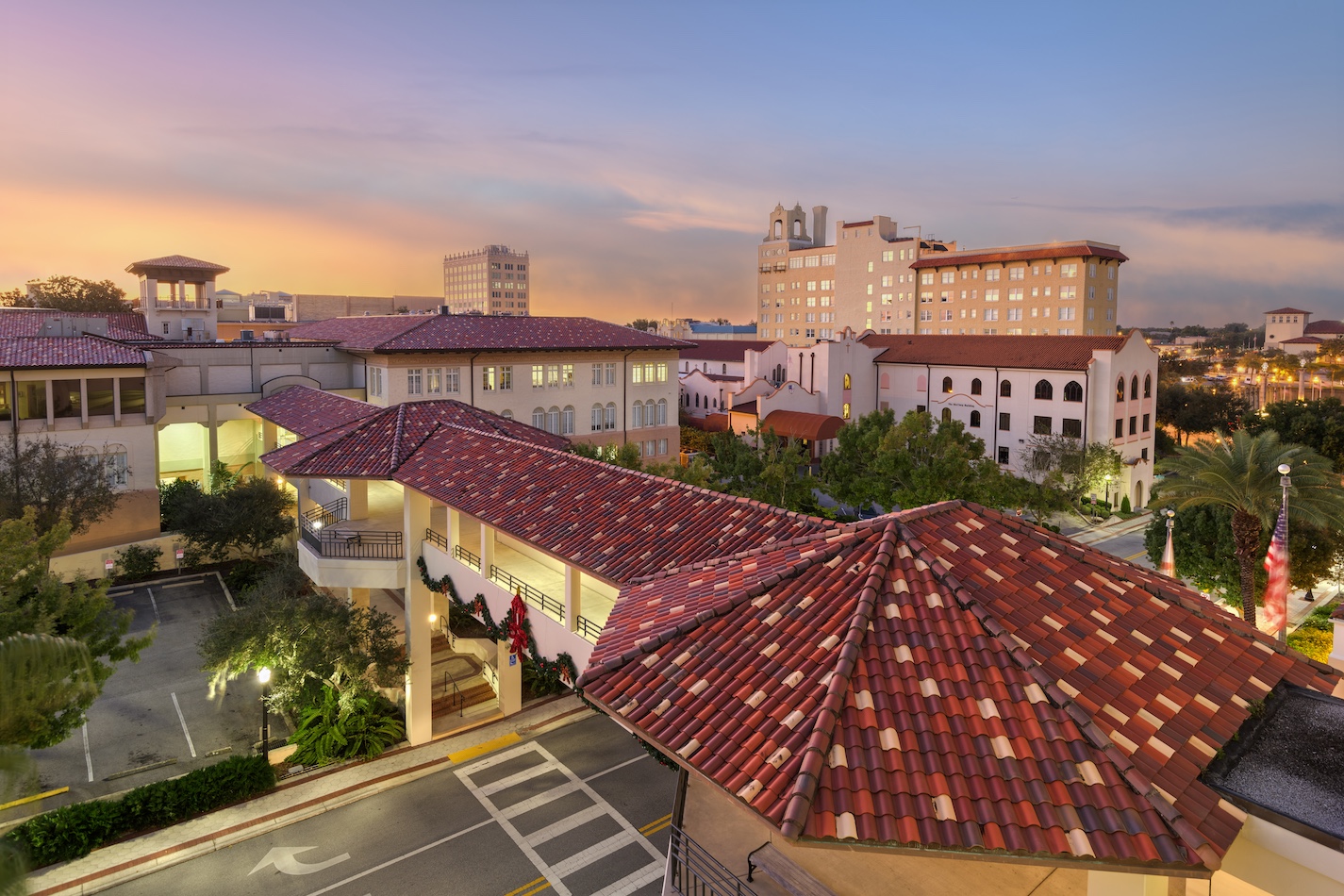 An elevated view of a picturesque Mediterranean-style campus at sunset. Red-tiled roofs and off-white walls dominate the scene, with palm trees dotting the campus. Recently pressure washed, the buildings gleam against the soft, colorful evening sky in the background.