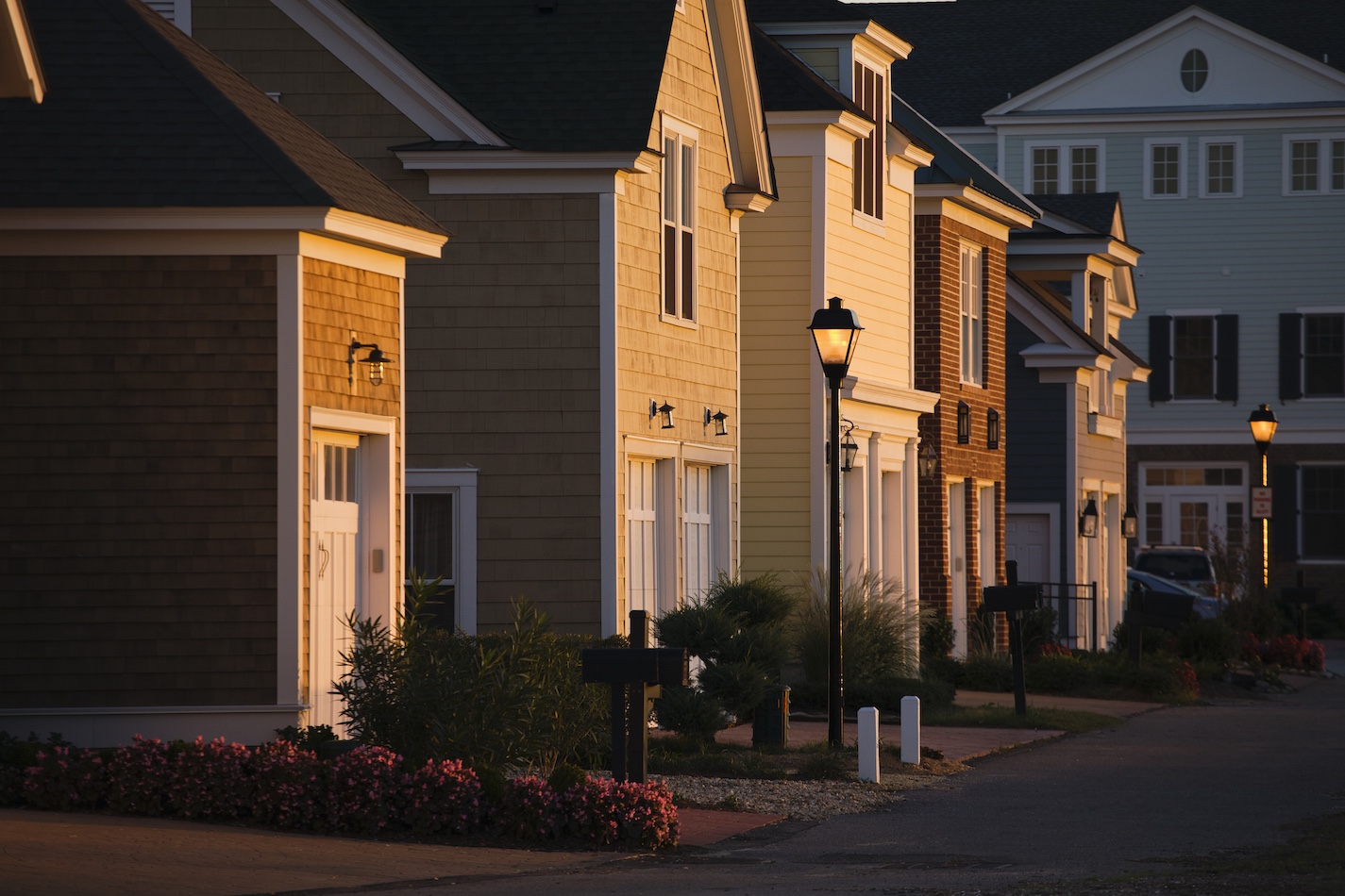 Rows of suburban houses with various shades of beige and blue siding, lit warmly by the evening sun. The street, lined with lampposts and small, well-manicured gardens, showcases the community's attention to detail and excellent services. A larger building can be seen in the background.