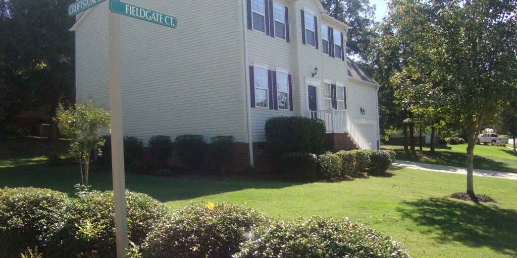 A two-story, light-colored house with black shutters, surrounded by a well-manicured lawn and shrubs. On the corner is a signpost for "Coopman Ct" and "Fieldgate Ct." The sky is clear and blue, with trees lining the background.