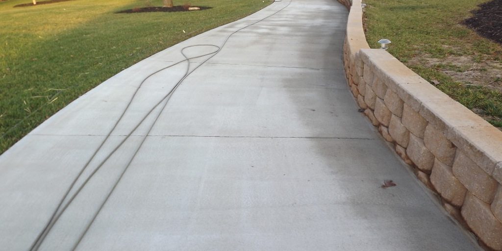 A wide, newly paved concrete driveway, perfect for pressure washing, leads up to a large brick house with multiple windows and a red front door. The driveway is bordered by a low stone retaining wall on one side. The surrounding area is landscaped with grass and small trees.