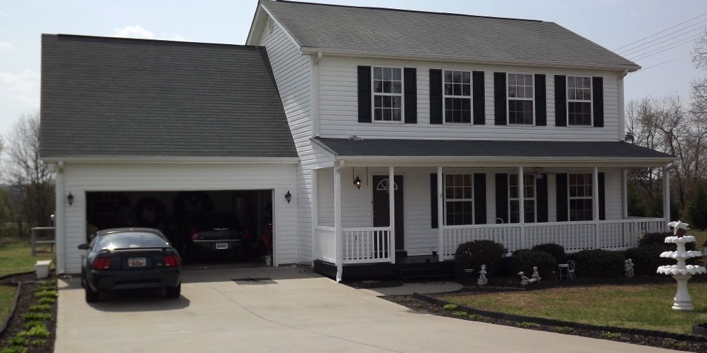 A two-story white house with black shutters and a gray roof. There's a front porch with columns and a double garage on the left. A black car is parked in the driveway, which has been recently pressure washed. The yard features minimal landscaping and a white decorative structure on the right.