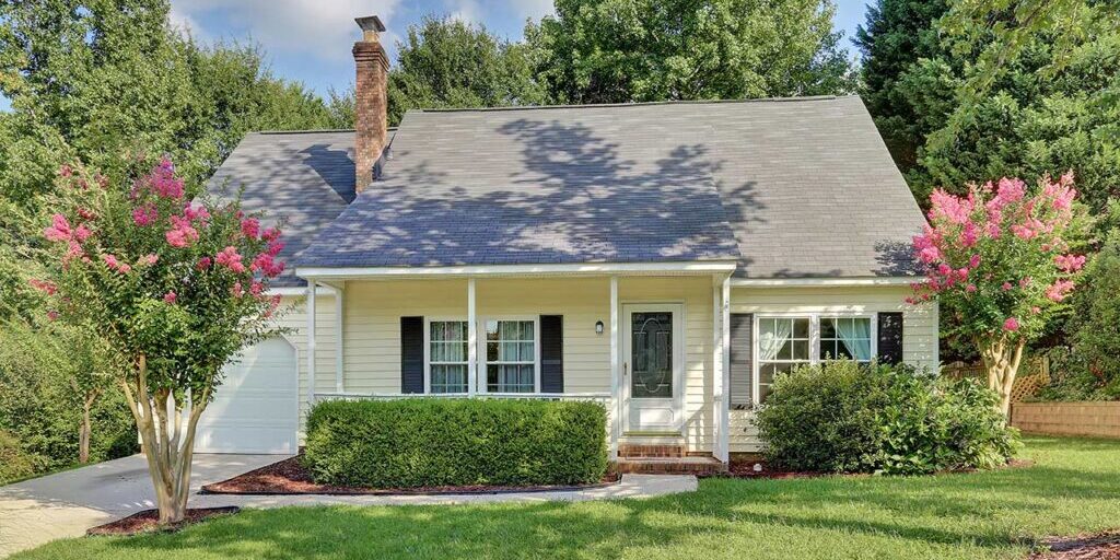 A charming single-story house with light-colored siding, a gray roof, and a covered front porch. The front yard includes two flowering trees, well-maintained shrubs, and a patch of green grass. The house has a driveway leading to an attached garage.