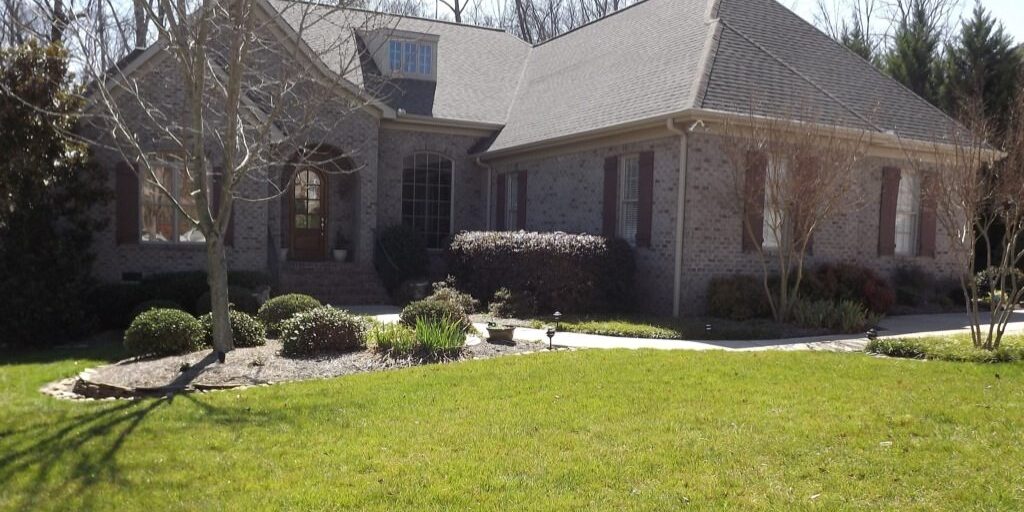 A single-story brick house with a large, well-manicured front lawn and sparse landscaping including bushes and small trees. The house, which has recently undergone pressure washing, boasts a gabled roof and a front entrance with steps leading up to it. Leafless trees are visible in the background.