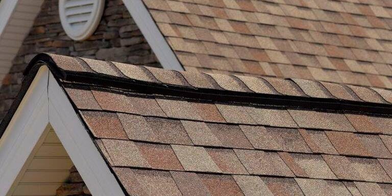 A close-up view of the rooftops of a house with brown and beige composite shingles, white trim, and a gable vent. The exterior walls are covered with stone siding, and the sky in the background is clear with a few clouds.