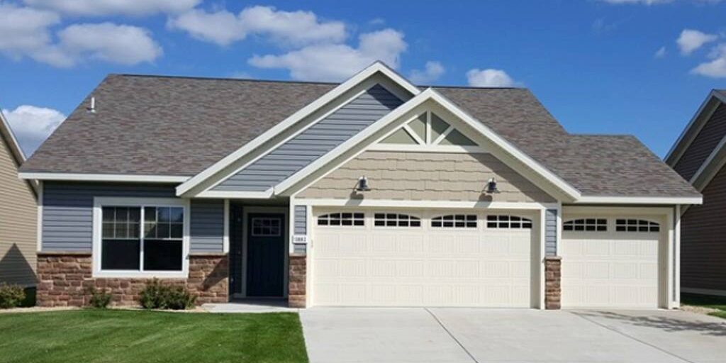 A single-story house with gray and beige exterior, dark blue front door, and three-car garage. The driveway and sidewalk are made of concrete, and the front yard features a well-maintained green lawn. The sky above is clear with a few white clouds.