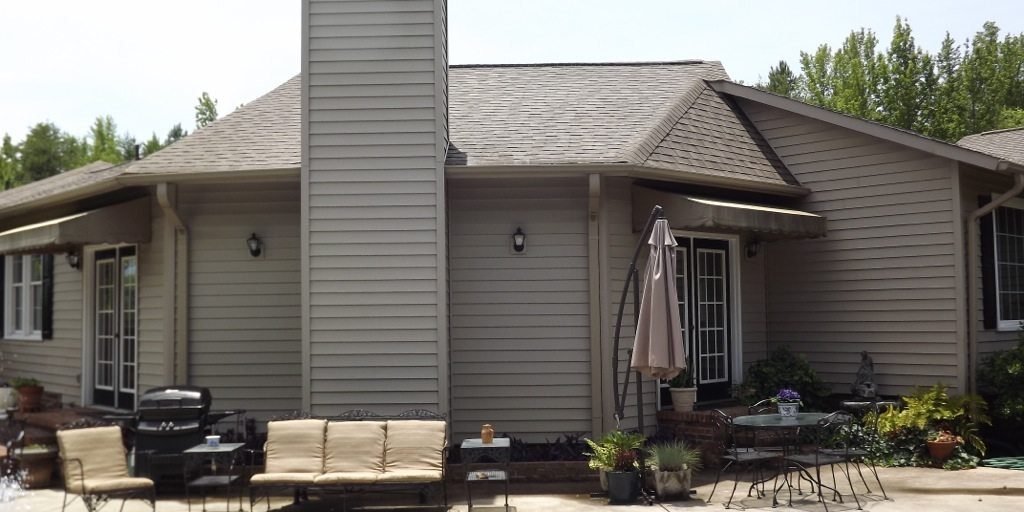 A one-story house with beige siding and a large central chimney. The backyard features patio furniture, including a beige couch and chairs, a black grill, and a table with an umbrella. In the foreground, there is a blue swimming pool. Recently pressure washed, trees are visible in the background.