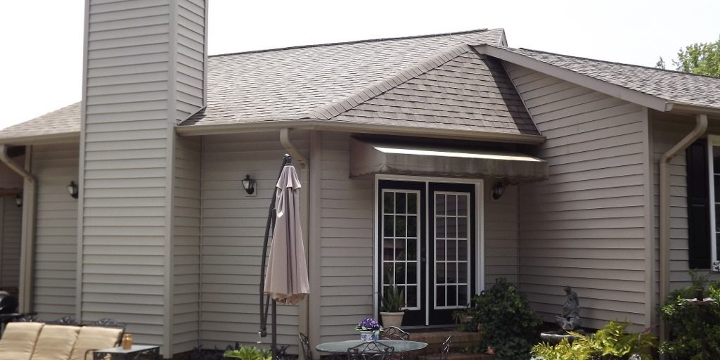 A single-story house with beige siding features a central chimney, double French doors with glass panes, and an awning above the doors. Recently pressure washed, the patio in the foreground includes a table with chairs, potted plants, and an outdoor umbrella.