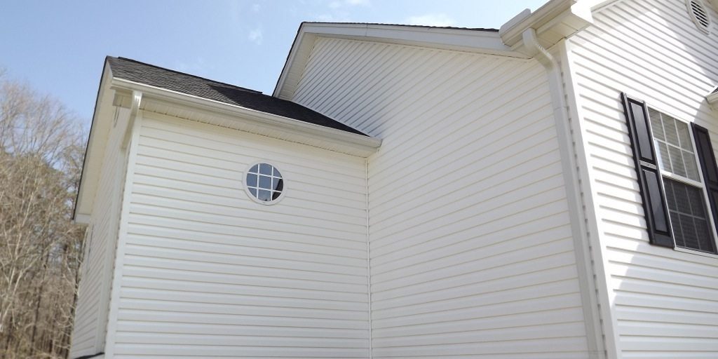 A close-up of the exterior of a white house. The house features clean, white horizontal siding, black shutters on the windows, and a circular window on the smaller section of the building. The base has a gray concrete foundation, suggesting recent pressure washing. The sky is clear with some wispy clouds.