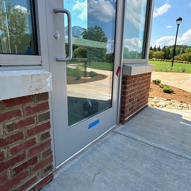 A glass entrance door to the commercial building is framed by a metal door frame and brick wall. A blue tape is seen near the base of the door. Behind the glass, part of an interior space with a panel and a sign are visible. Outside, there is a sidewalk and green landscape, possibly prepped for pressure washing.