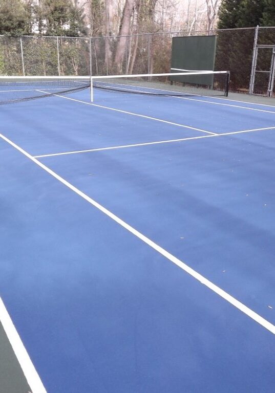 A tennis court with a blue playing surface and green outer areas. The net is set up in the middle, and the court is surrounded by a chain-link fence with trees and foliage in the background. Recently undergoing pressure washing, it looks pristine and ready for play.