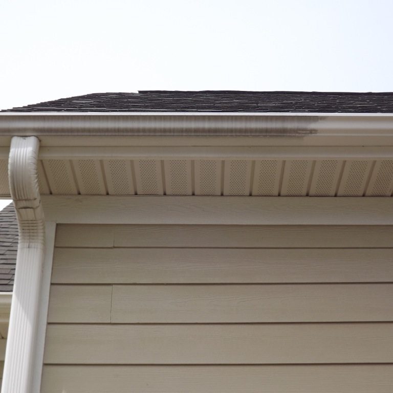 A house roof with beige vertical siding is shown. The image captures the gutter system, with a downspout attached to the side of the house. The roof appears to have dark shingles and a slight overhang. There's a noticeable dark streak on the gutter, likely needing pressure washing for a clean finish.