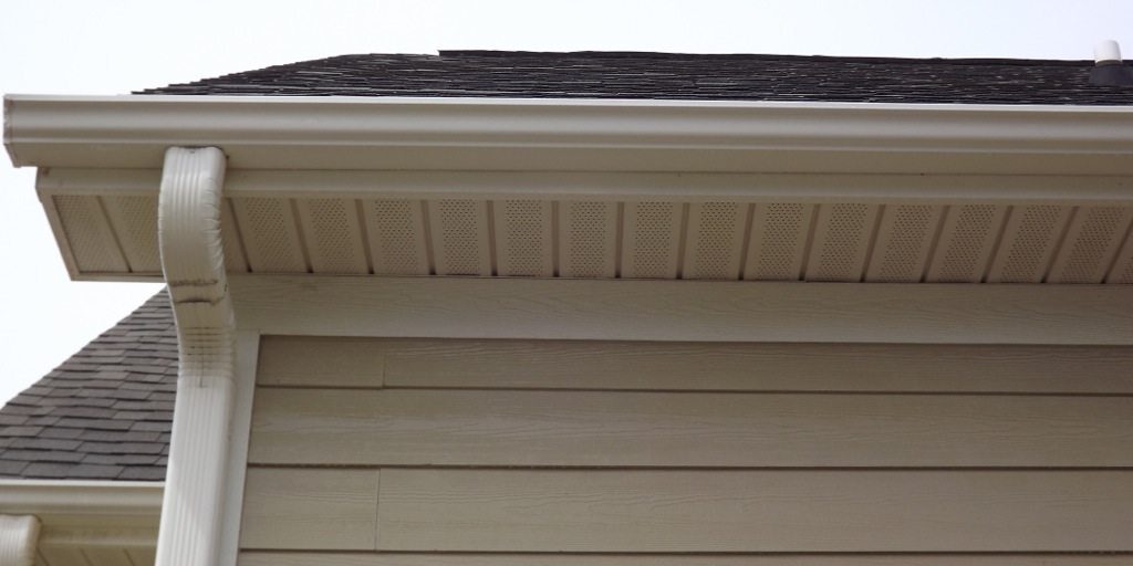 The image shows the side of a house with beige siding and a white rain gutter system, ideal for maintaining through regular pressure washing. The roof has dark shingles, and the soffit features ventilation holes. The gutter downspout is visible running down the corner of the house. The sky is clear.