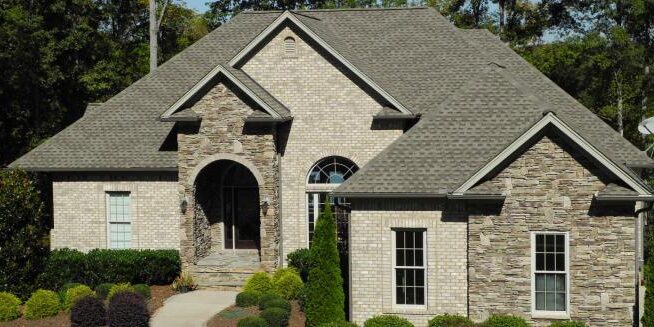 A two-story house with a combination of brick and stone exterior. The entrance features an arched doorway and front steps. The roof is peaked and shingled. The house is surrounded by green shrubs and trees.