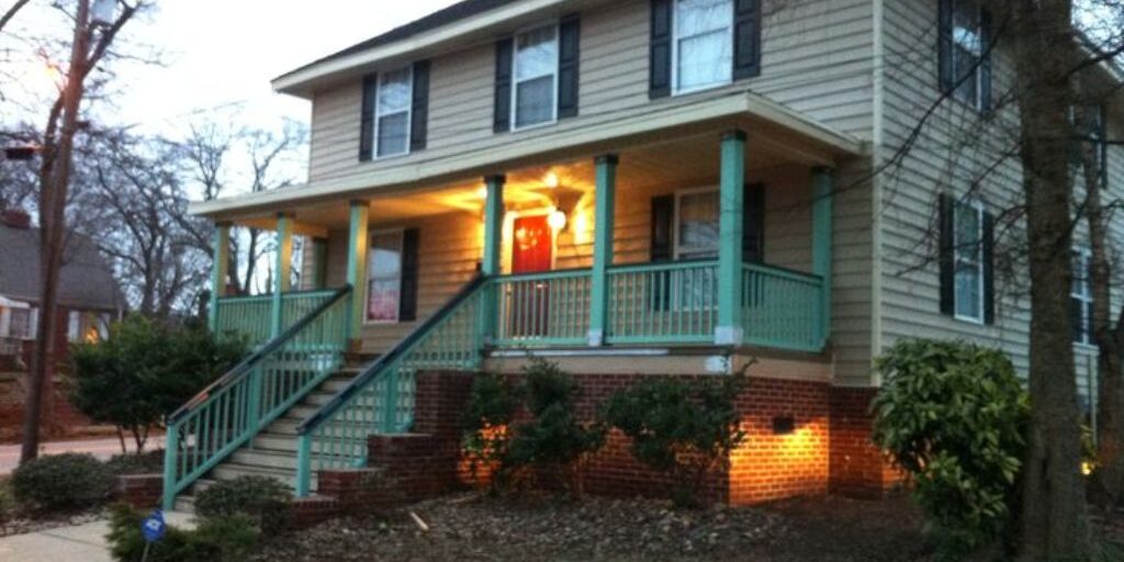 A two-story house with beige siding and teal trim, featuring a red front door with a lit porch light. The home has a spacious front porch, brick foundation, matching teal steps, and bushes surrounding parts of the house. The setting appears to be around dusk.