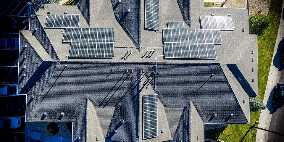 An aerial view of a residential rooftop with multiple solar panels installed. The roof has a complex structure with various angles and ridges. Surrounding the house, there are driveways, pathways, and landscaped areas visible.