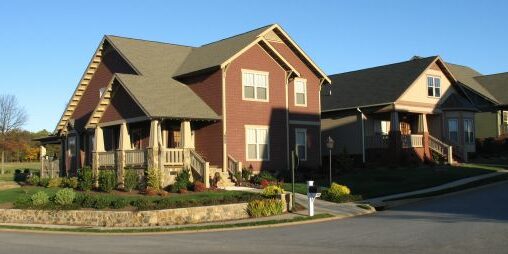 A suburban street corner featuring two single-family homes with porches. The house on the left is maroon with beige trim, and the house on the right is beige. Both have well-maintained yards with shrubs and flowers. The sky is clear and blue.