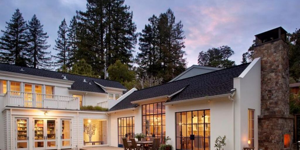 A modern two-story house with large windows and outdoor seating area. The ground level features a dining table with chairs on a patio, while the upper level has a balcony. Tall trees surround the house, and the sky is slightly cloudy, creating a serene atmosphere.