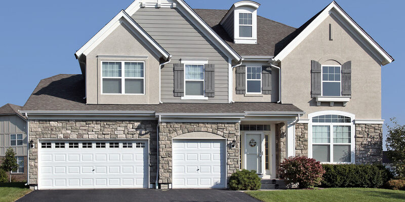 A two-story house with a combination of stone and siding exterior. The house has a symmetrical design with two garage doors, shuttered windows, and a small porch. The lawn is well-kept with bushes and small trees. The sky is clear and blue.