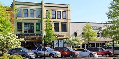 A quaint downtown street scene showing a row of various colorful commercial buildings including shops and businesses. Several cars are parked along the street, and trees with green foliage line the road, adding a touch of nature to the urban setting.