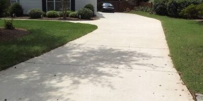 A concrete driveway curves slightly to the right leading up to a garage attached to a house. The driveway is flanked by well-kept green grass and shrubs. Shadows of trees are cast on the driveway. The house has light-colored siding, and a car is parked in the driveway.