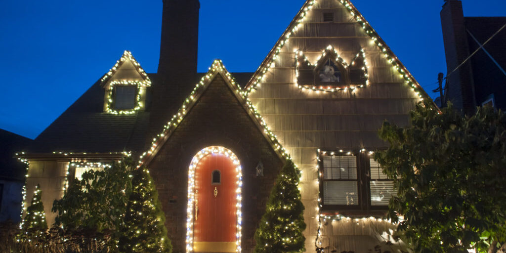 A quaint house with a pointed roof decorated with white Christmas lights. The house features a prominent chimney, arched red door, and surrounding bushes also adorned with lights. The sky is dusky blue, indicating early evening.