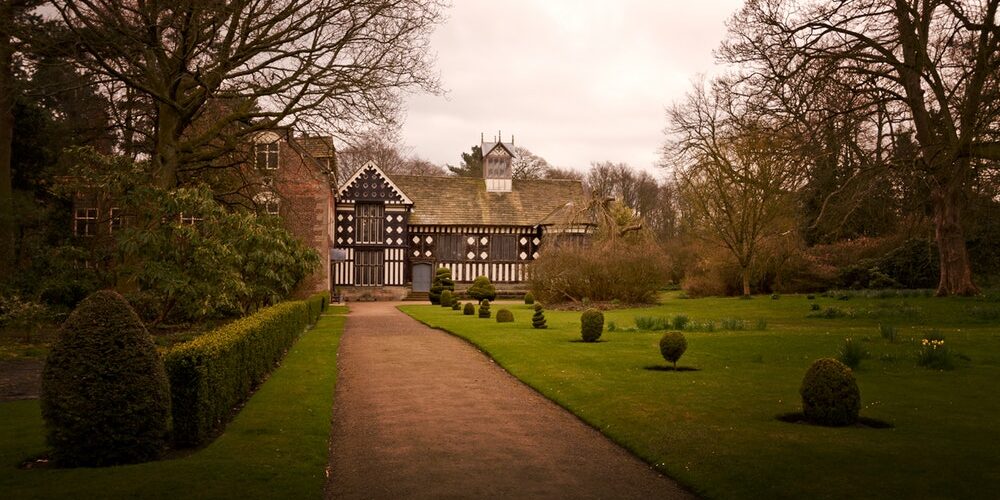 A historic Tudor-style house stands at the end of a dirt path, flanked by manicured hedges and topiary. Leafless trees surround the scene, with cloudy skies overhead. The checkered black and white facade of the house contrasts with its green lawn and shrubs.